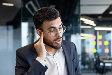 Mature man businessman with glasses experiencing ear pain, possibly due to inflammation or otitis, located in a modern corporate office. The professional attire and modern office background