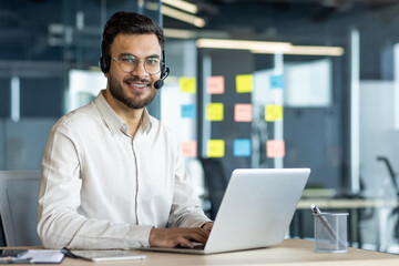 A mature male business professional, wearing glasses and a headset, types on a laptop in an office. The workspace exemplifies a collaborative and productive corporate environment