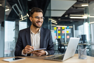 Fototapeta premium Mature professional businessman engaged in an online webinar in a modern office environment. Taking notes on a notepad while using a laptop, he displays attentiveness and focus.