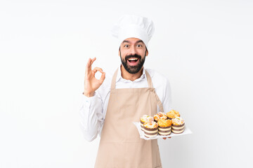 Young man holding muffin cake over isolated white background surprised and showing ok sign