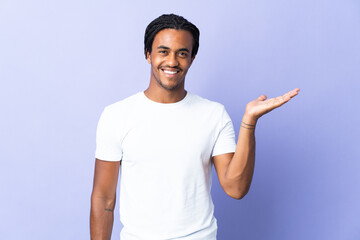 Young African American man with braids man isolated on purple background holding copyspace imaginary on the palm to insert an ad