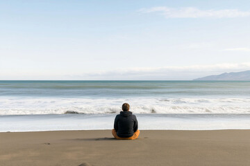 Burnout recovery stress recharge and relaxation. A person sits on a beach, gazing at the ocean waves under a clear sky, embodying tranquility and reflection.