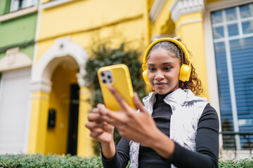 Young latin woman using smartphone and wearing yellow headphones outdoors