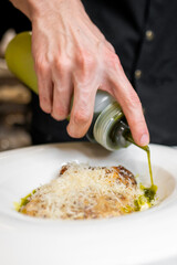 A close-up of a chef's hand drizzling green oil over a plated dish topped with grated cheese. The focus is on the gourmet presentation and meticulous attention to detail in the kitchen.