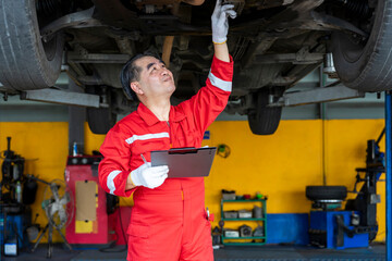 asian senior experienced mechanic business owner in uniform checking lifted vehicle in garage...