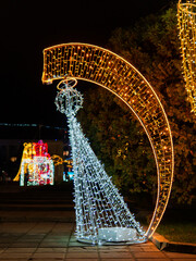 Colorful holiday lights create a vibrant display in a park at night. A white tree structure stands next to a large golden arch, adding to the festive atmosphere that celebrates the season.