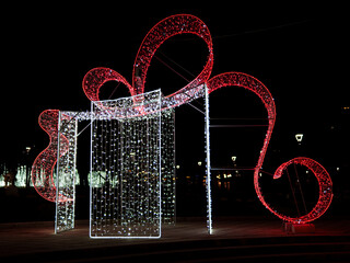A vibrant holiday light display features a large gift shape adorned with red and white lights. The installation shines brightly against the night sky, creating a festive atmosphere in a public space.