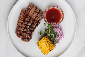 Overhead view of a delicious grilled striploin steak with grilled corn on the cob, pickled red onions, and a small bowl of barbecue sauce, placed on tabletop. Restaurant or cafe menu, cookbook recipe