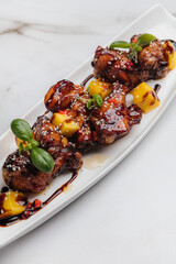 Glazed chicken wings are served on a white rectangular plate, garnished with mango chunks, sesame seeds, fresh basil leaves, and a drizzle of sauce. Restaurant or cafe menu, cookbook recipe