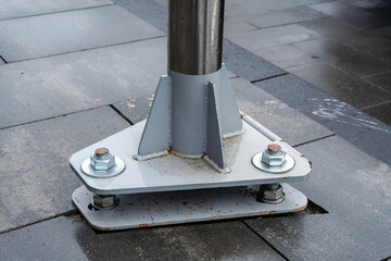 A sturdy metal pole base is firmly anchored with bolts to a stone pavement in a city environment under natural light