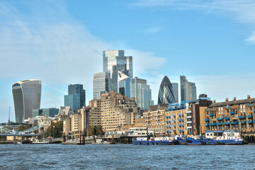 Fototapeta premium Modern London skyline featuring iconic skyscrapers like the Gherkin and Walkie Talkie, viewed from the River Thames. Historic buildings, boats, and blue skies. London.