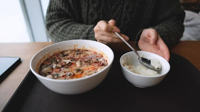 Pretty brunette enjoying delicious Tom Yum Kun soup with shrimps in Thai restaurant winter in Siberia. A woman in a warm cozy sweater is having lunch near the window. Snow outside