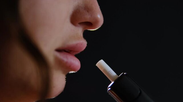 Woman vaping an electronic cigarette, close-up of lips exhaling vapor against a dark background, emphasizing smoking habits.