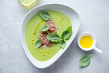Bowl of cold green pea soup topped with prosciutto and basil, horizontal shot on a light-grey granite surface, high angle view