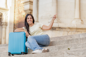 Young woman has just arrived in the city with her travel suitcase and points to the side to show a monument or the way to the hotel.