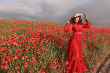 A girl in a red dress and hat stands in a field of poppy flowers at sunset