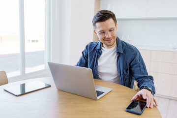 Man working on laptop at bright desk while using smartphone in a modern living space