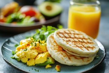 A delicious breakfast plate featuring scrambled eggs, avocado, arepas, and a glass of juice.