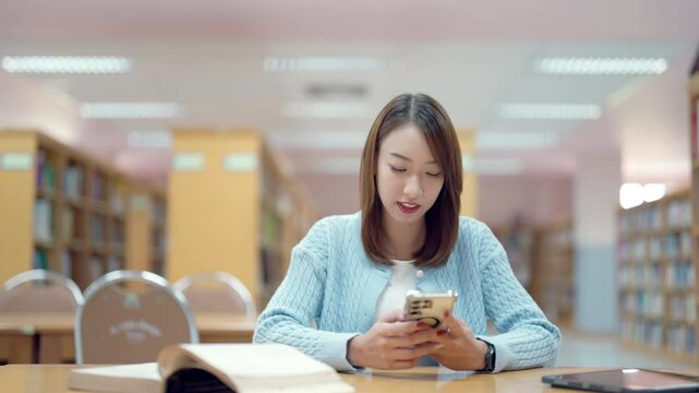 A woman is sitting at a table in a library, looking at her cell phone. She is wearing a blue sweater and she is focused on her phone. The scene suggests a quiet and studious atmosphere