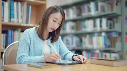 A woman is sitting at a table in a library, using a tablet to read. She is focused and engaged in her work