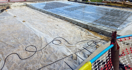 construction site with a concrete foundation being prepared. The area includes a large section of plastic sheeting covering the ground, with steel reinforcement bars (rebar) 