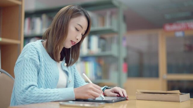 A woman is sitting at a table in a library, writing on a tablet. She is wearing a blue sweater and she is focused on her work. The scene suggests a quiet and studious atmosphere
