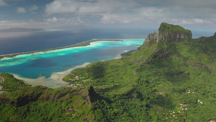 Breathtaking aerial view of Bora Bora island, French Polynesia, vibrant turquoise lagoon, lush green vegetation, and majestic mount Otemanu under a cloudy sky. Wild travel destination, remote nature