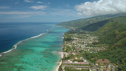Tahiti island cityscape high angle view, French Polynesia. Vibrant coral reef, turquoise waters, lush green mountains, and coastal settlements village, houses, road under bright blue sky. Drone shot
