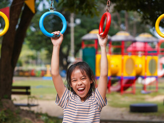 Happy girl hanging on monkey bar by hand doing exercise. Little Asian girl playing at outdoor playground in the park on summer vacation. Healthy activity.