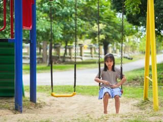 A sad little girl is sitting alone on a playground swing. An unhappy child is playing alone.