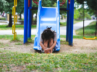 A sad little girl is sitting alone on a playground slide. An unhappy child is playing alone.