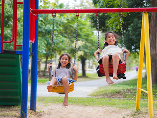 Cute sisters are having fun playing on the swings in a playground in a park. Children playing in the playground during summer vacation.