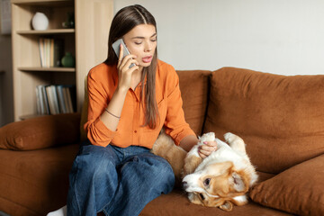 Young lady worried about her sick Corgi dog calling vet, sitting on couch and touching pet's paw