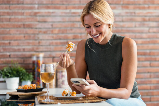 Pretty young woman eating sushi while using smartphone in the kitchen at home - Powered by Adobe