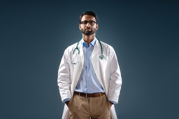 Serious, pensive Indian doctor, bearded man wearing white coat, stylish eyeglasses, hands in pockets