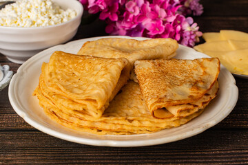 Pancakes are on a white plate, there is a bowl of sour cream next to it, a plate of cheese and cottage cheese on a wooden table