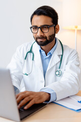 Indian professional doctor wearing white coat, stylish eyeglasses, using laptop, typing on keyboard