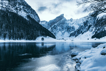 Frozen alpine lake with black ice surrounded by snow-covered mountains that reflects on the ice,...