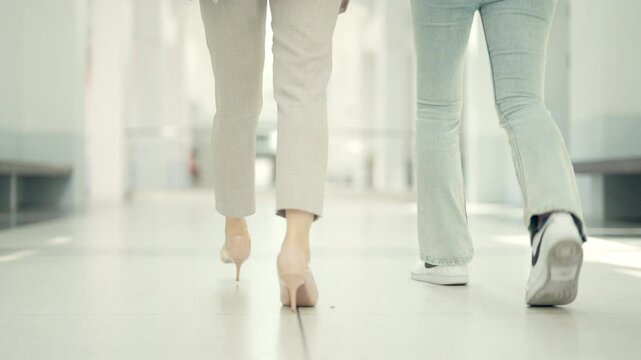 Two women walking down a hallway in a hospital. One is wearing high heels and the other is wearing sneakers
