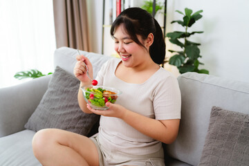 Beautiful Asian woman eating salad, holding a glass bowl and using a fork to pick up a tomato to eat