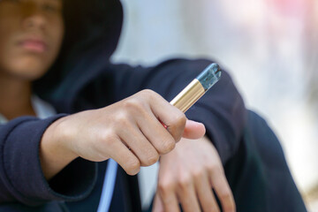 Close-up of hand holding out electronic cigarette, blurred background of a teenage boy wearing black hoodie over his head.