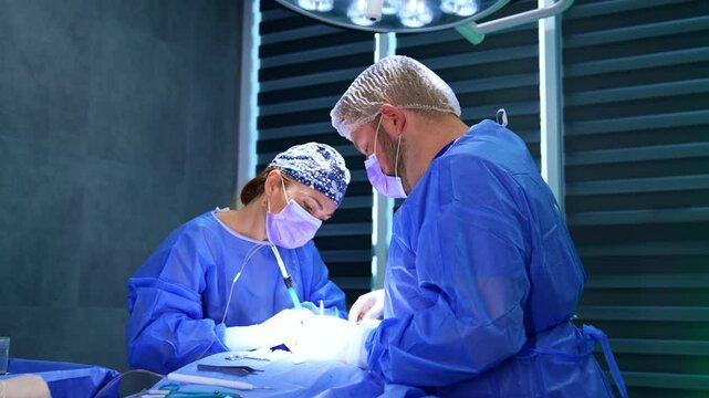 Female surgeon and assisting male medic work at operation. Doctors work under the bright round lamp.