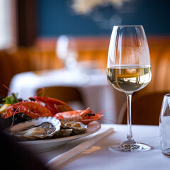 A photo of a close-up shot of a table in a cozy restaurant. There is a glass of white wine in the foreground, next to a plate of appetizing seafood