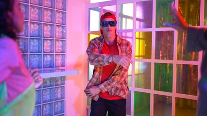 Caucasian dancer dancing while diverse friend looking at footstep in neon light at night club. Back view of multiethnic hipster moving to freestyle music with happy man looking at camera. Regalement.