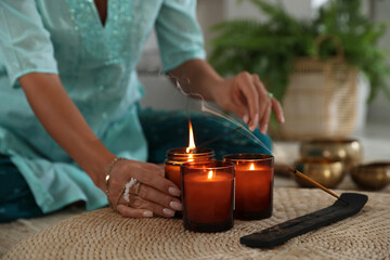 Woman lighting incense sticks with burning candles indoors, closeup