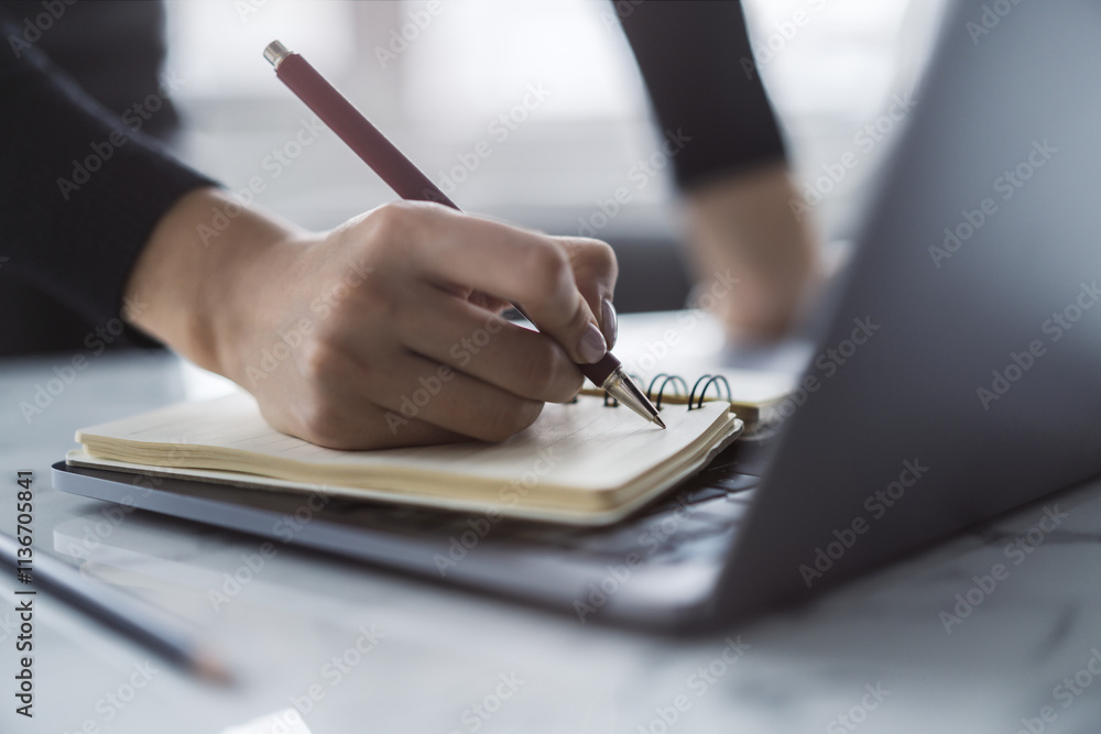 Poster The image captures a woman's hand in close view as it writes in a notepad on a chic laptop, background out of focus