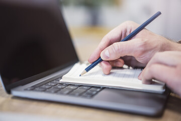 Focused view of a man's hand as he writes in a notepad on a modern laptop, background gently blurred