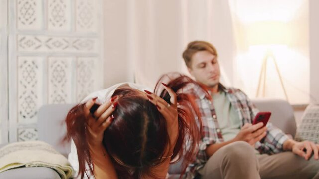 A multiethnic couple sits on a couch at home. The wife looks upset, while the husband appears indifferent, focused on his phone instead of their conversation.