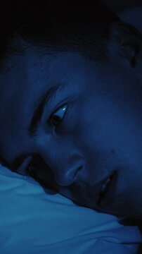 Vertical video. Nightmare fear. Sleep disorder. Disturbed scared frightened guy woke up opened eyes in bed late at night in dark blue light.
