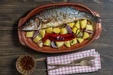 Delicious baked big sea bass fish with spices in a clay plate and fried vegetables, closeup, top view. Cooked sea bass with potatoes, red chili pepper, onion and garlic